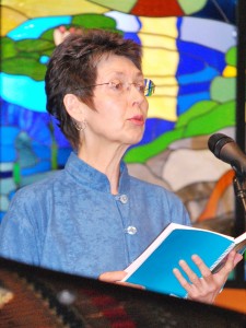 Head and upper-torso view of a woman with short brown hair, holding a music book and singing into a microphone