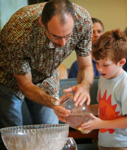 Man assists a boy to pour water from a vase into a larger bowl