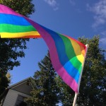 Rainbow flag billowing against a background of blue sky, tree foliage and part of a house