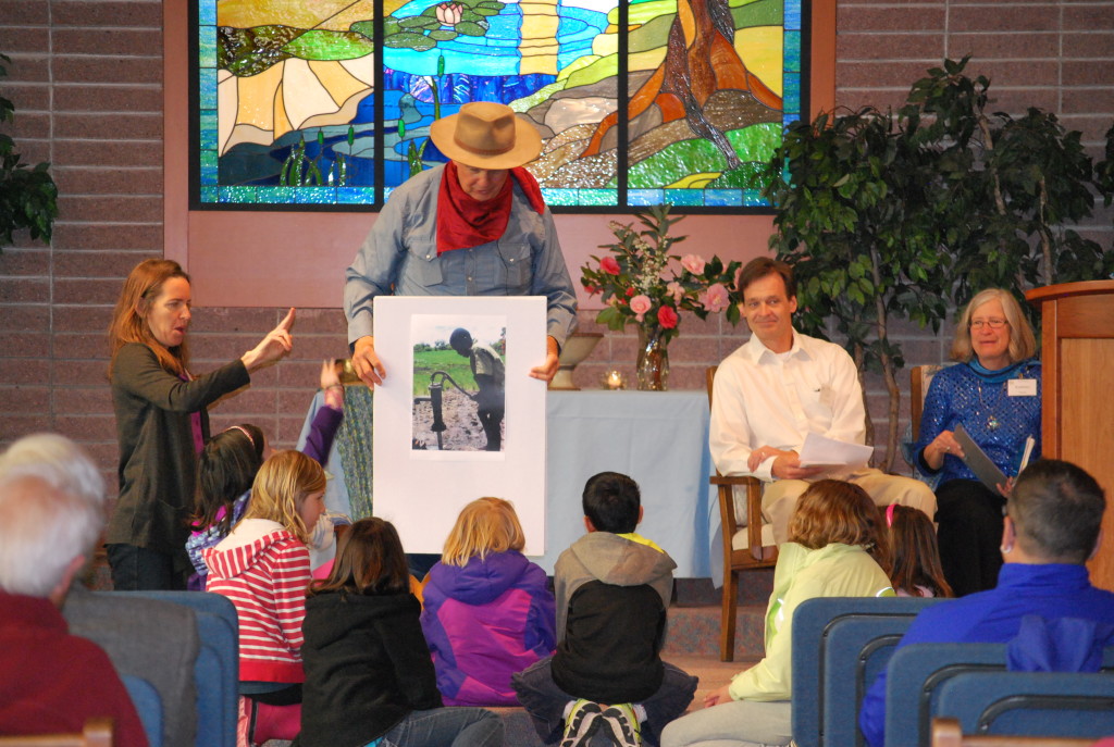 Mike Evans, dressed in a farmer's costume, displays a photograph of a person priming an old-fashioned water pump while, at Mike Evans' feet, children look on