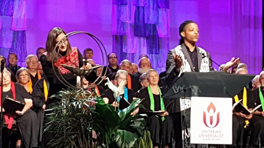 A woman at left lights UUA General Assembly chalice while another woman speaks at a lectern, right. Behind them, a group of people look on, wearing black garments with colorful stoles