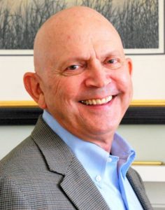 Head-and-shoulders photo of a smiling, beige-skinned man wearing a collared light-blue shirt and a gray suit-jacket