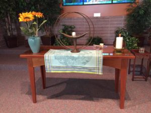 RVUUF's chalice, a brass-colored basin with candle surrounded by double interlocking circles, sits atop white cloth on a wooden table, flanked by a vase of sunflowers on one side and a wide and tall white candle on the other side