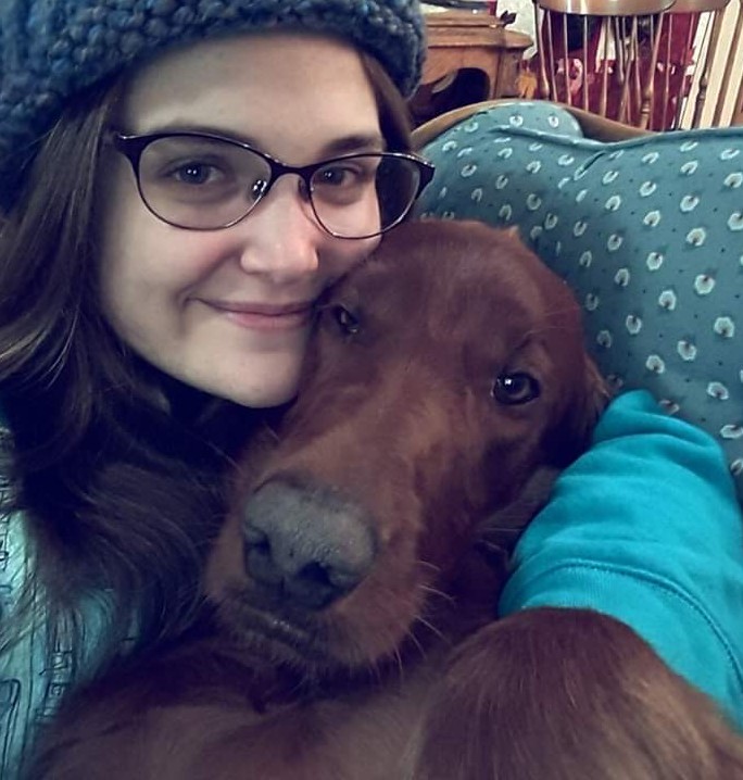 Close-up photo of a beige-skinned, dark-haired woman snuggling a reddish-brown dog on blue sofa