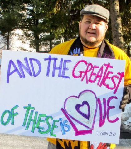 Rev. Sean smiling and holding a hand-lettered sign saying "And the greatest of these is Love"